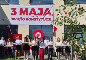 Uczennica wcielająca się w rolę Konstytucji zaprasza uczniów na słodki poczęstunek, na scenie także dziewczynki trzymające tace z babeczkami