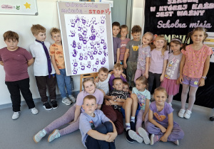 uczniowie klasy 1a prezentujący wspólnie wykonane zadanie – odbite fioletowe rączki na tablicy