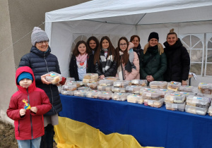 Grupa uczniów, nauczycieli sprzedających ciasto oraz rodzic z dzieckiem, kupujący ciasto.